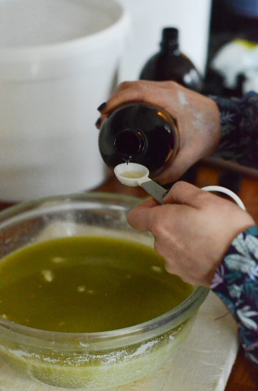 A person measuring and pouring liquid from a dark amber bottle into a spoon over a glass bowl filled with green mixture, likely during a skincare or soap-making process.