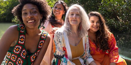 A diverse group of four smiling women of different ages, including an older woman with grey hair, sitting outdoors in natural sunlight, wearing colourful, bohemian-style clothing and enjoying each other’s company in a lush green setting.