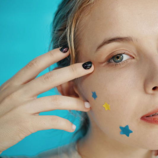 Close-up of a young woman with star-shaped acne patches on her cheek, touching her face with manicured fingers against a blue background