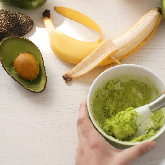 A bowl of mashed banana and avocado being mixed by hand, surrounded by a peeled banana and halved avocado on a light wooden surface, showing ingredients for a natural DIY face mask.