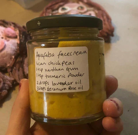 A glass jar of homemade aquafaba face cream with a handwritten label listing ingredients including chickpeas, xanthan gum, turmeric powder, lavender oil, and geranium rose oil.