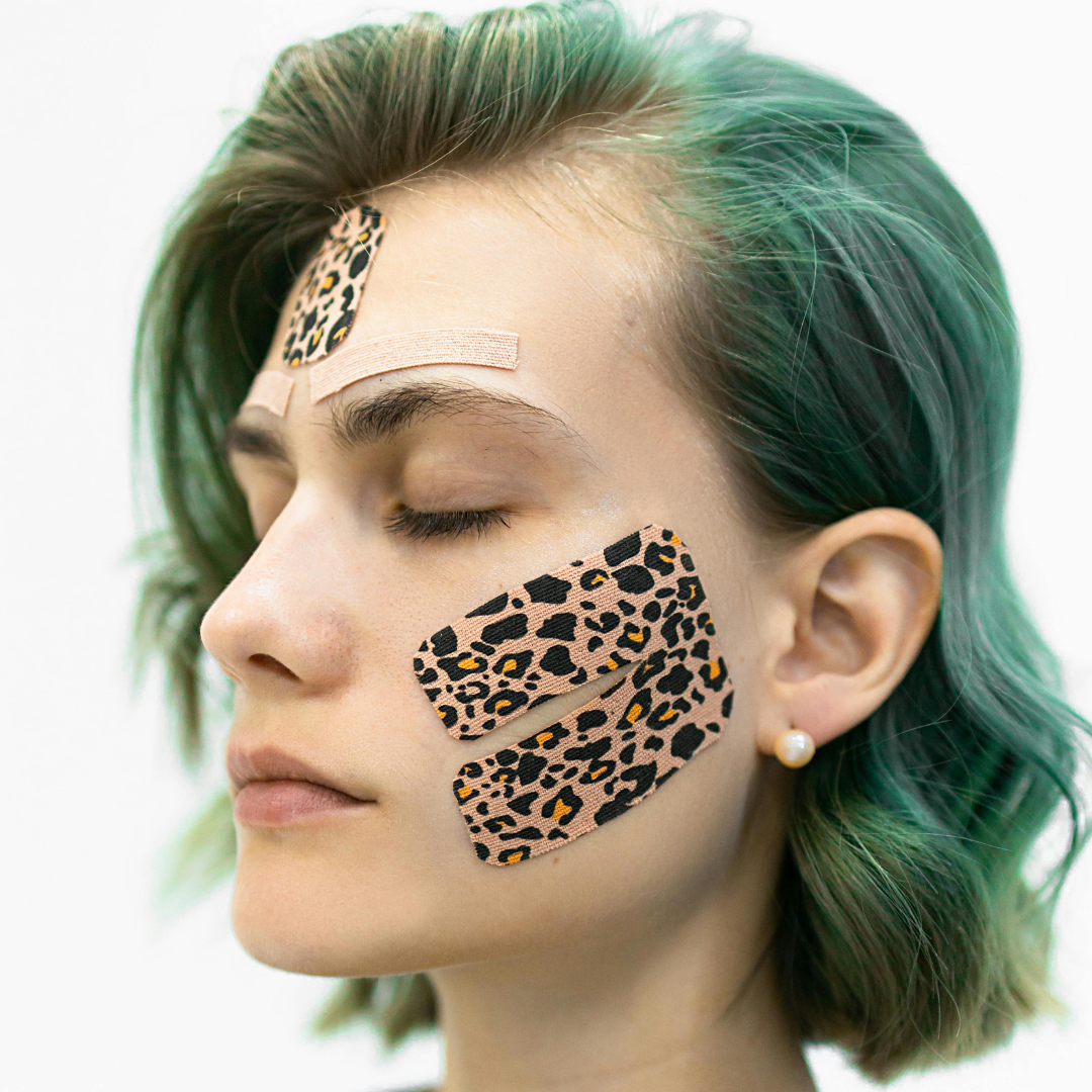 Young woman with green hair wearing leopard-print skin tape patches on her forehead and cheeks, eyes closed