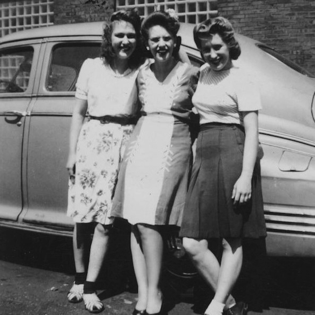 Three women standing arm in arm and smiling in front of a vintage car, wearing mid-20th-century dresses and skirts, in a black-and-white photograph.