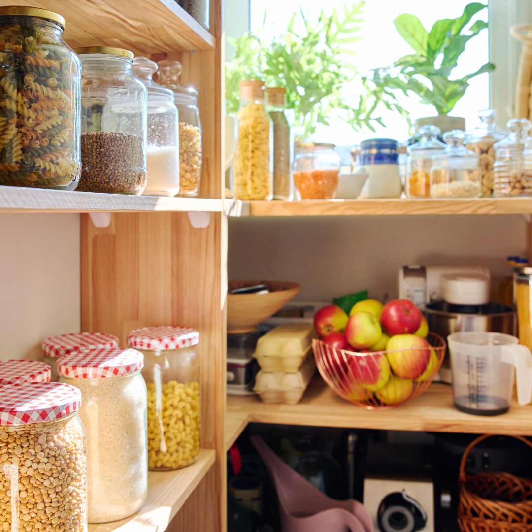 A bright kitchen pantry filled with glass jars of pasta, grains and pulses, with a bowl of apples and natural light streaming through green plants on the windowsill