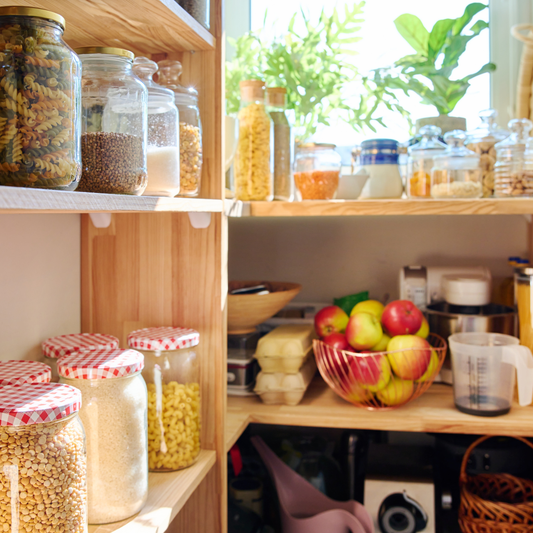 A bright kitchen pantry filled with glass jars of pasta, grains and pulses, with a bowl of apples and natural light streaming through green plants on the windowsill