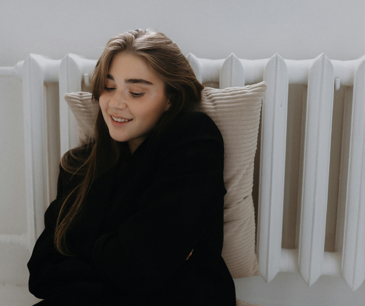 Young woman with long brown hair sitting comfortably against a cushion and white radiator, smiling softly while looking down, wearing a black jumper in a calm, cosy setting.