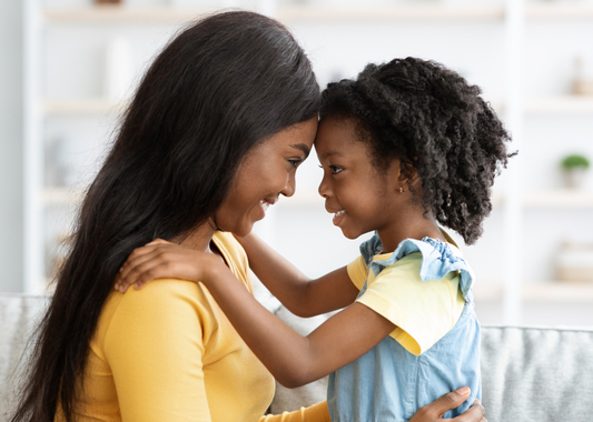 black women with long hair, looking into a young black girls eyes, their foreheads are touching and the young girl has her hands on the women's shoulders