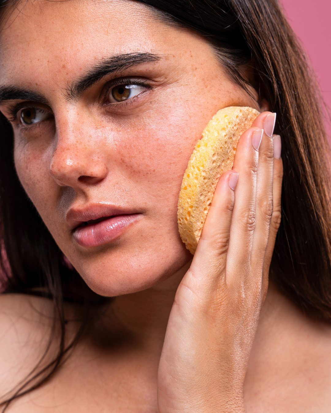 women with dark hair, eyebrows and eyes, holds a round yellow sponge to her cheek
