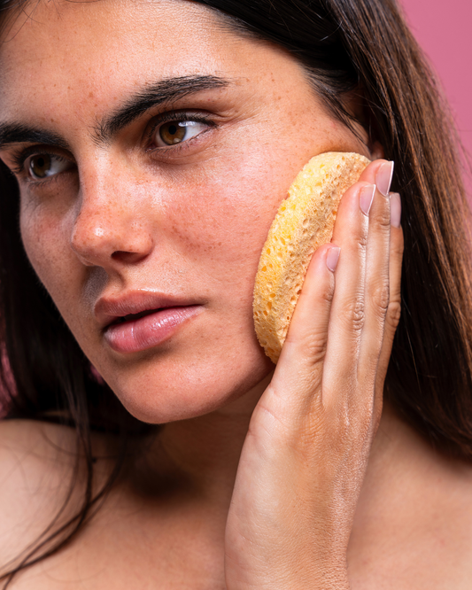 women with dark hair, eyebrows and eyes, holds a round yellow sponge to her cheek