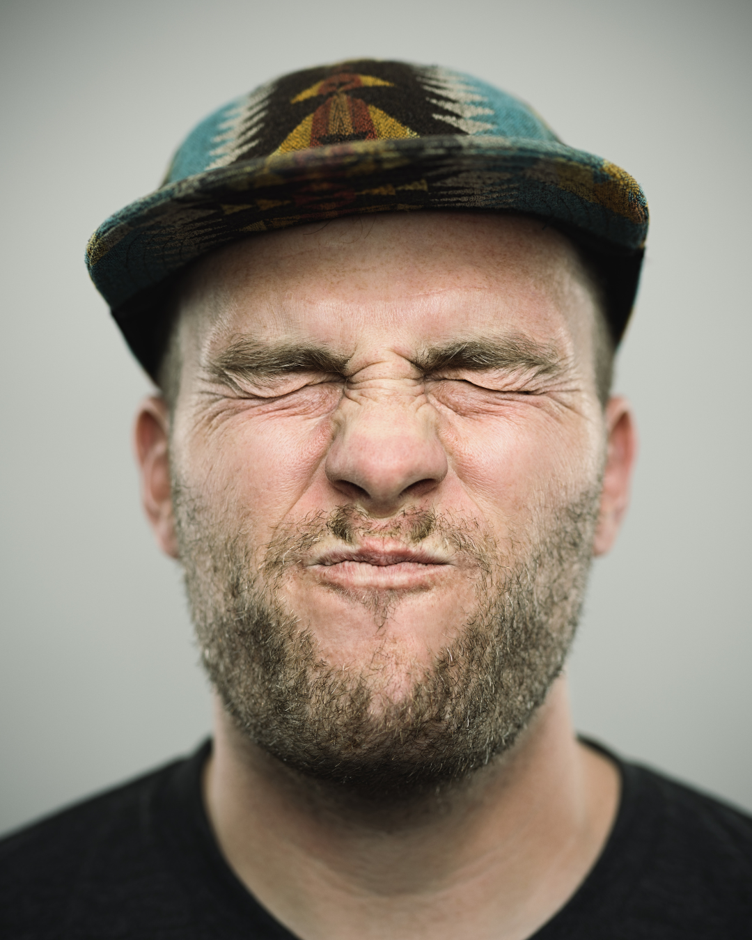 Close-up of a man with a beard wearing a patterned cap, eyes closed and face scrunched, showing skin texture and expression